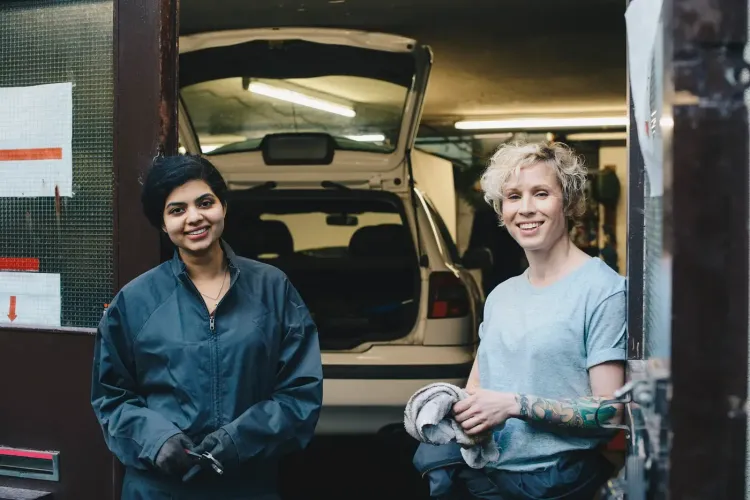 Two smiling women in work attire stand in a garage with an open car trunk.