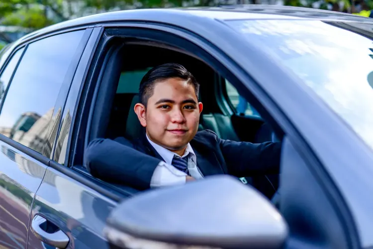 Young man in a suit sitting in a gray car, looking at the camera.
