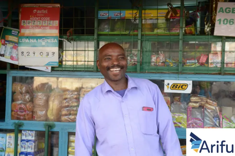 A smiling man with a mustache stands in front of a shop displaying packaged goods.