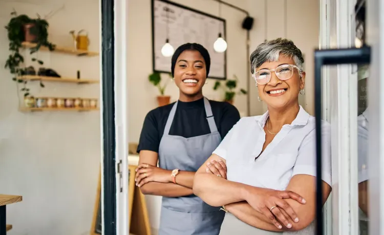 Two smiling women, an older owner and younger employee, stand proudly in their cafe doorway.
