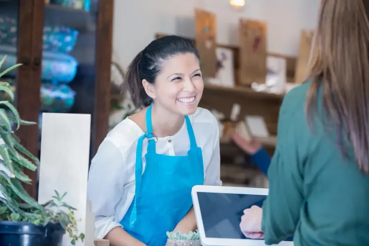 Smiling female cashier in blue apron assists a customer at a bright counter.