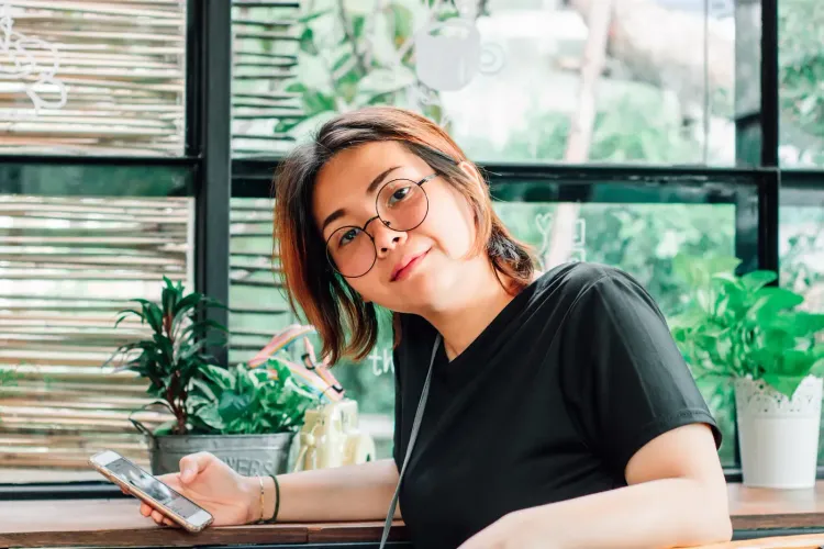 Woman with glasses holding a phone, smiling at the camera in a cafe setting.