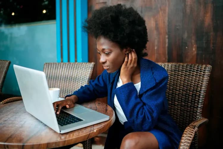 Black woman in a blue blazer typing on a laptop in a cafe setting.