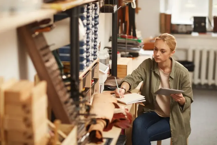 Young woman working intently in a leather workshop, writing notes and using a tablet.