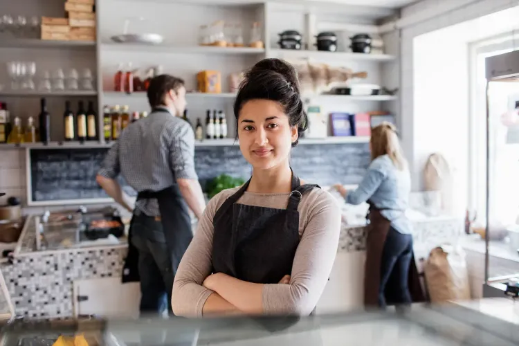 Smiling woman in an apron with crossed arms stands confidently in a busy cafe.