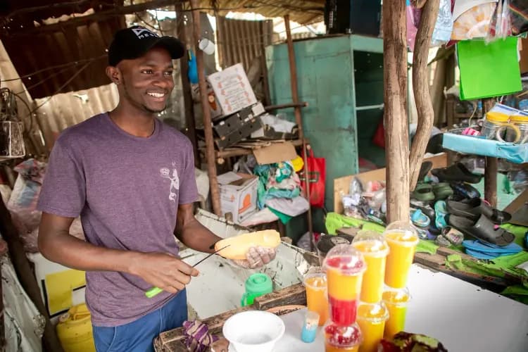 Smiling man at a street stall selling colorful layered drinks and fresh food.