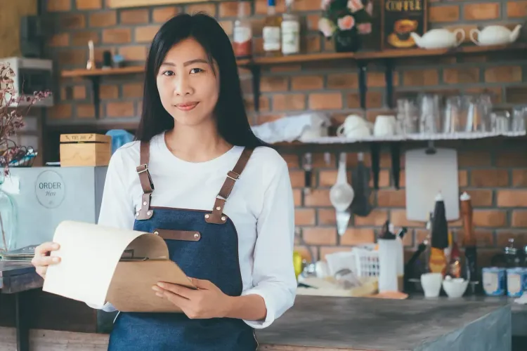 A young Asian woman in a cafe apron holds a menu, smiling at the viewer.