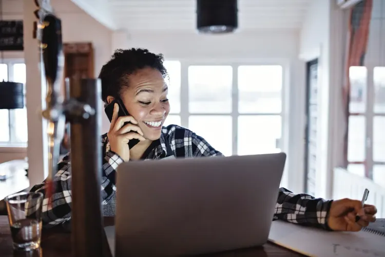 Smiling woman on phone, working on laptop at a bar counter.