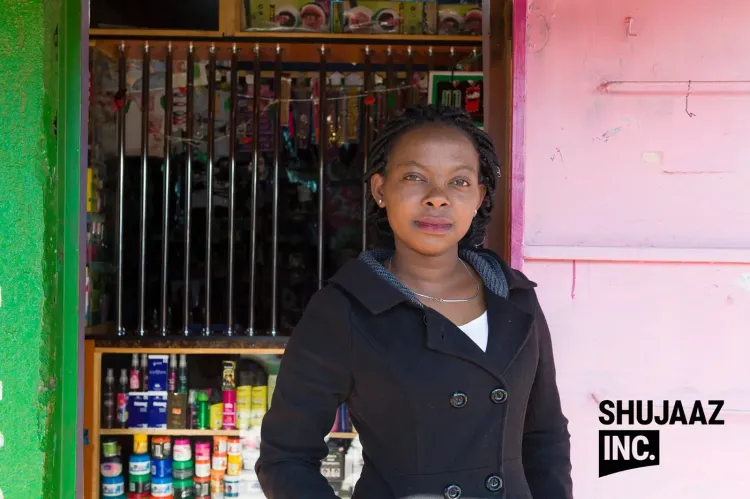A woman stands in front of her small retail shop displaying various products.
