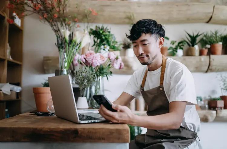 A man in an apron uses his phone and laptop in a flower shop.