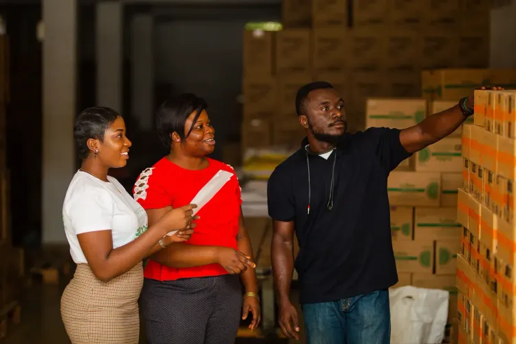 Three people in a warehouse reviewing inventory, with a man pointing at stacked boxes.