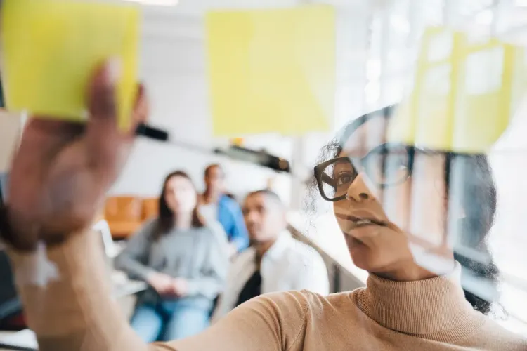 Woman in glasses puts a yellow sticky note on a clear board during a team meeting.