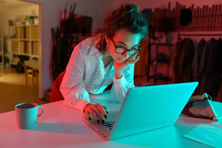 Woman in glasses intently working on a laptop bathed in vibrant red and blue light.