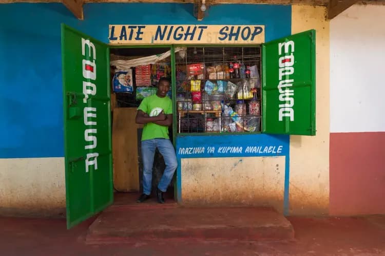 A man in a green shirt stands in the open doorway of a small "Late Night Shop".
