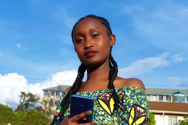 Young woman with braids in a patterned top holds a phone under a blue sky.