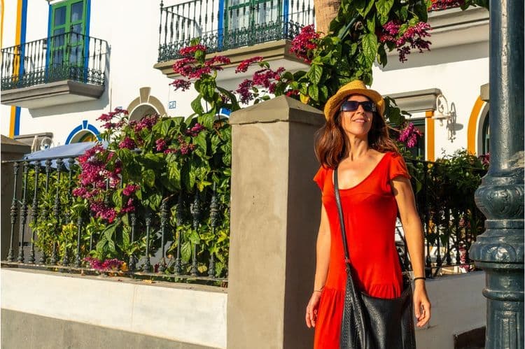 Smiling woman in a red top and straw hat walks past colorful buildings with flowers.
