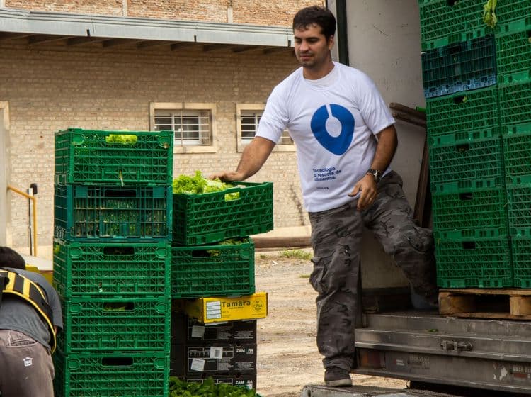 A man in a white shirt with a logo moves green crates filled with fresh produce.