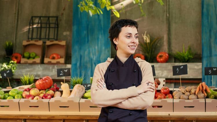 Young market vendor smiling with arms crossed, surrounded by fresh produce.