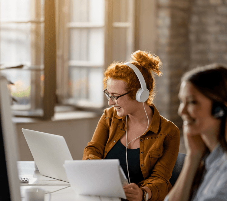 Two smiling women with headphones work on laptops in a modern office.
