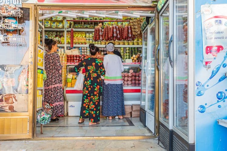 Three women shop inside a small, brightly lit market stocked with various goods.