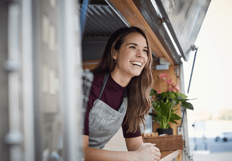 A young woman wearing a gray apron smiles, looking out from her food truck window.