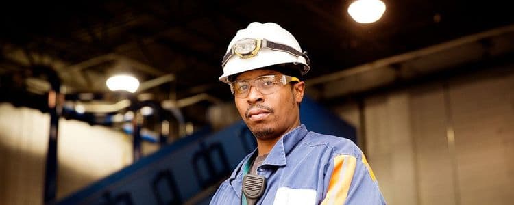 Man in hard hat, safety glasses, and work uniform in an industrial setting.