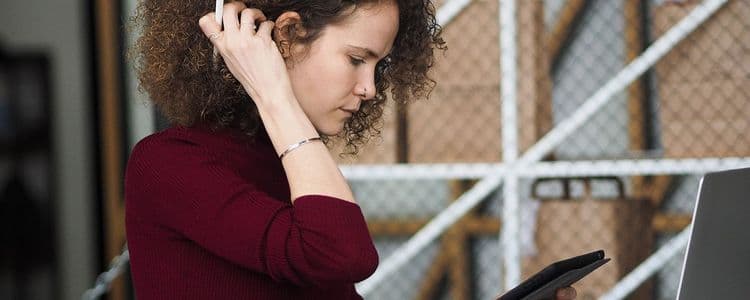 Curly-haired woman in maroon shirt looking intently at a tablet, hand near her head.