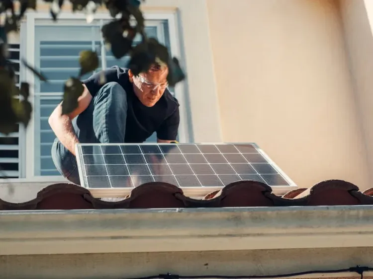 A man installs a solar panel on a red tiled residential roof under sunlight.
