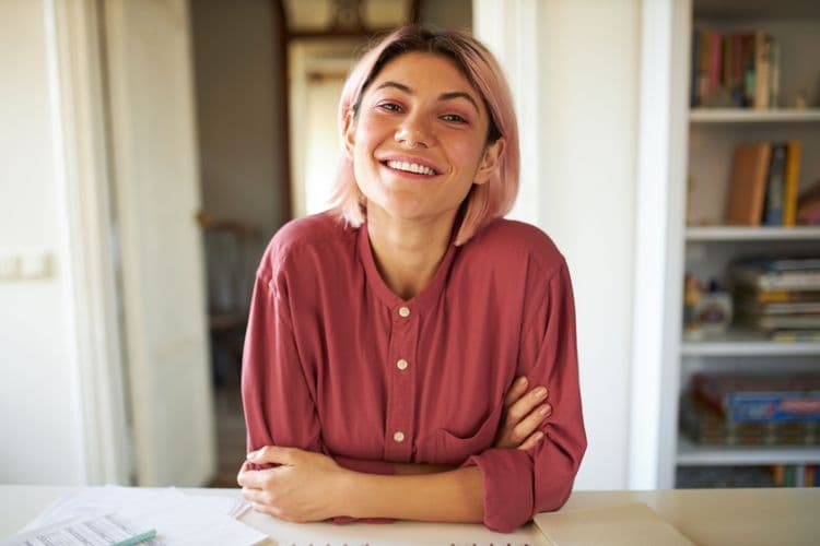 Young woman with pink hair smiling brightly at the camera, wearing a red shirt.