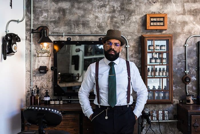 Man with beard, fedora, and suspenders stands confidently in a rustic barbershop.
