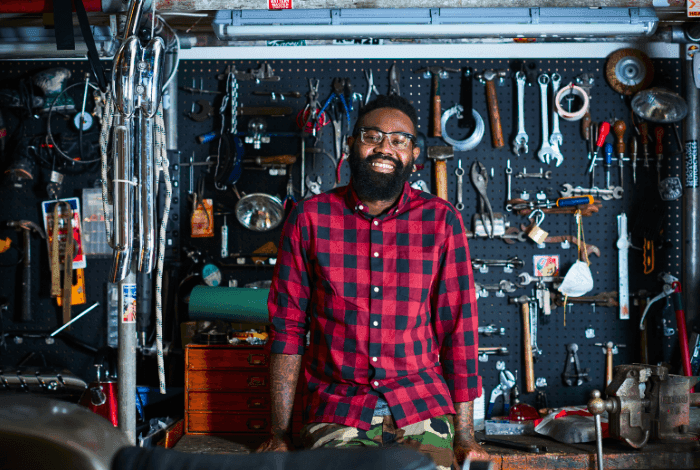 Bearded man in plaid shirt smiling in a cluttered workshop with tools on a pegboard.